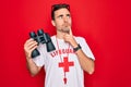 Young handsome lifeguard man wearing t-shirt with red cross and whistle using binoculars serious face thinking about question,