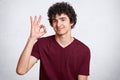 Young handsome male with curly hair, makes okay sign, shows his approval, gestures with hand, isolated over white background. Sati Royalty Free Stock Photo