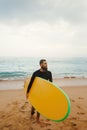 Young handsome male surfer with a surfboard is enjoying a view while walking a sandy beach at sea. Summer, vacation, ocean.