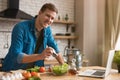 Young handsome man adding spices to vegetables while preparing salad for lunch standing in well-equipped modern kitchen checking
