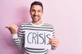 Young handsome man asking for economy holding paper with crisis word message pointing thumb up to the side smiling happy with open