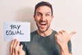 Young handsome man asking for equality economy holding paper with pay equal message pointing thumb up to the side smiling happy