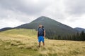 Young handsome man with backpack standing in mountain grassy valley on copy space background of summer woody mountain peak and Royalty Free Stock Photo