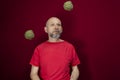 Young handsome man with beard, bald head and red shirt standing tossing pinecone fruit up against red background