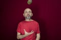 Young handsome man with beard, bald head and red shirt standing tossing pinecone fruit up against red background