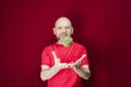 Young handsome man with beard, bald head and red shirt standing tossing pinecone fruit up against red background