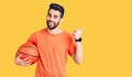 Young handsome man with beard hoilding basketball ball pointing thumb up to the side smiling happy with open mouth Royalty Free Stock Photo