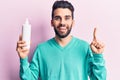 Young handsome man with beard holding bottle of cream smiling with an idea or question pointing finger with happy face, number one Royalty Free Stock Photo