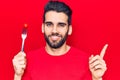 Young handsome man with beard holding fork with tomato smiling happy pointing with hand and finger to the side