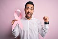 Young handsome man with beard holding pink cancer ribbon over isolated background screaming proud and celebrating victory and Royalty Free Stock Photo