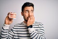 Young handsome man with beard holding plastic denture teeth over white background looking stressed and nervous with hands on mouth