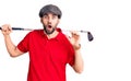 Young handsome man with beard playing golf holding club and ball scared and amazed with open mouth for surprise, disbelief face