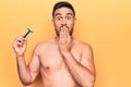 Young handsome man with beard shirtless holding depilation razor over yellow background covering mouth with hand, shocked and