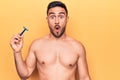 Young handsome man with beard shirtless holding depilation razor over yellow background scared and amazed with open mouth for