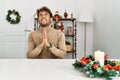 Young handsome man with beard sitting on the table by christmas decoration begging and praying with hands together with hope Royalty Free Stock Photo