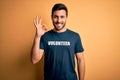Young handsome man with beard volunteering wearing t-shirt with volunteer message doing ok sign with fingers, excellent symbol