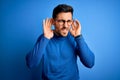 Young handsome man with beard wearing casual sweater and glasses over blue background Trying to hear both hands on ear gesture, Royalty Free Stock Photo