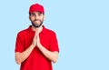 Young handsome man with beard wearing delivery uniform praying with hands together asking for forgiveness smiling confident