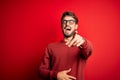 Young handsome man with beard wearing glasses and sweater standing over red background laughing at you, pointing finger to the Royalty Free Stock Photo