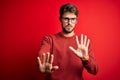 Young handsome man with beard wearing glasses and sweater standing over red background Moving away hands palms showing refusal and Royalty Free Stock Photo