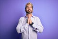 Young handsome man with beard wearing golden crown of king over purple background begging and praying with hands together with