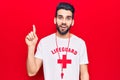 Young handsome man with beard wearing lifeguard t-shirt and whistle smiling with an idea or question pointing finger up with happy