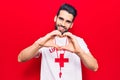 Young handsome man with beard wearing lifeguard t-shirt and whistle smiling in love doing heart symbol shape with hands