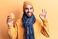 Young handsome man with beard wearing scarf and wool cap drinking orange juice doing ok sign with fingers, smiling friendly Royalty Free Stock Photo