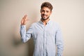 Young handsome man with beard wearing striped shirt standing over white background Waiving saying hello happy and smiling, Royalty Free Stock Photo