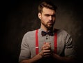 Young handsome man with beard wearing suspenders and bow tie, posing on dark background.