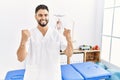 Young handsome man with beard working at pain recovery clinic very happy and excited doing winner gesture with arms raised, Royalty Free Stock Photo