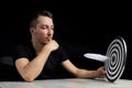 Man sits at table holding dart board with quill pen stuck in the center of the target on black background Royalty Free Stock Photo