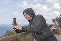 Young handsome man in black hood jacket photographs the sea with smartphone Royalty Free Stock Photo