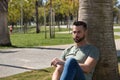 Young and handsome man, with blue eyes, beard, green t-shirt and jeans, sitting and leaning on a palm tree trunk and with a