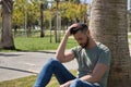 Young and handsome man, with blue eyes, beard, green t-shirt and jeans, sitting and leaning on the trunk of a palm tree, sad and Royalty Free Stock Photo