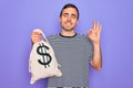 Young handsome man with blue eyes holding bag with dollar symbol over purple background doing ok sign with fingers, excellent Royalty Free Stock Photo