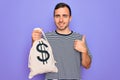 Young handsome man with blue eyes holding bag with dollar symbol over purple background happy with big smile doing ok sign, thumb Royalty Free Stock Photo