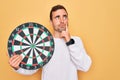 Young handsome man with blue eyes holding dartboard over isolated yellow background serious face thinking about question, very Royalty Free Stock Photo