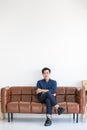 Young handsome man in blue shirt and jeans is sitting with his leg crossed at the middle on a big dark brown sofa with smile Royalty Free Stock Photo