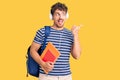 Young handsome man with curly hair holding student backpack and books pointing thumb up to the side smiling happy with open mouth Royalty Free Stock Photo