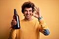 Young handsome man drinking bottle of beer standing over isolated yellow background doing ok sign with fingers, excellent symbol Royalty Free Stock Photo