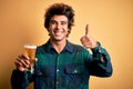 Young handsome man drinking glass of beer standing over isolated yellow background happy with big smile doing ok sign, thumb up Royalty Free Stock Photo