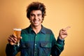 Young handsome man drinking glass of beer standing over isolated yellow background very happy pointing with hand and finger to the Royalty Free Stock Photo