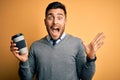 Young handsome man drinking a hot coffee from take away plastic cup over yellow background very happy and excited, winner Royalty Free Stock Photo