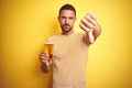 Young handsome man drinking a pint glass of beer over isolated yellow background with angry face, negative sign showing dislike Royalty Free Stock Photo