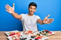 Young handsome man eating sushi sitting on the table looking at the camera smiling with open arms for hug Royalty Free Stock Photo