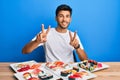 Young handsome man eating sushi sitting on the table smiling looking to the camera showing fingers doing victory sign Royalty Free Stock Photo