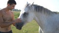 Young handsome man embracing and stroking white horse outdoors. Guy hugging a horse in the field, he caresses and