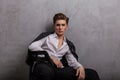 Young handsome man hipster in elegant white shirt in a fashionable leather black jacket is sitting on a chair near a gray vintage Royalty Free Stock Photo