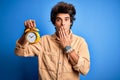Young handsome man holding alarm clock standing over isolated blue background cover mouth with hand shocked with shame for Royalty Free Stock Photo